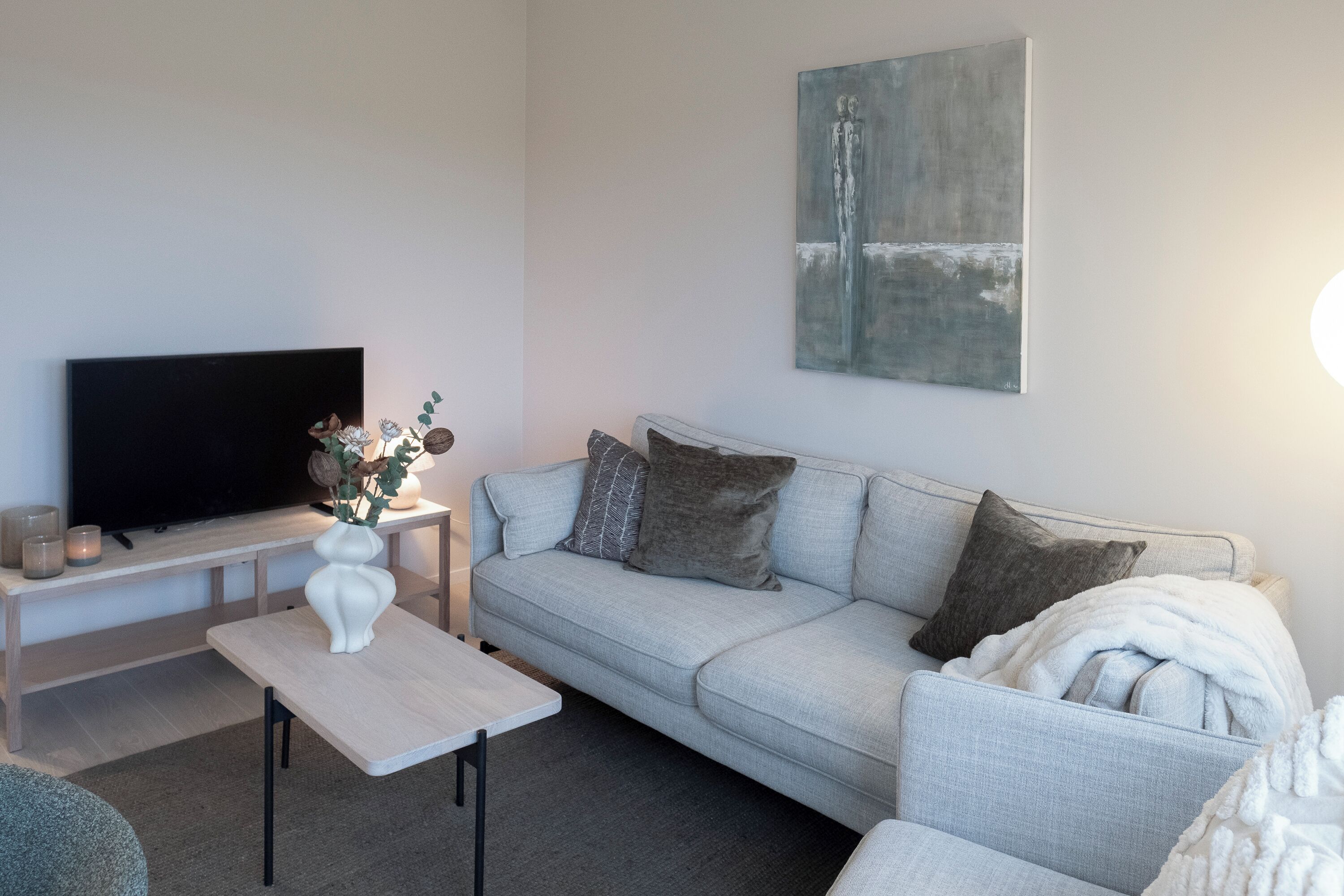 A modern living room with a grey sofa, a flat-screen TV, a white vase with flowers, and a painting on the wall.