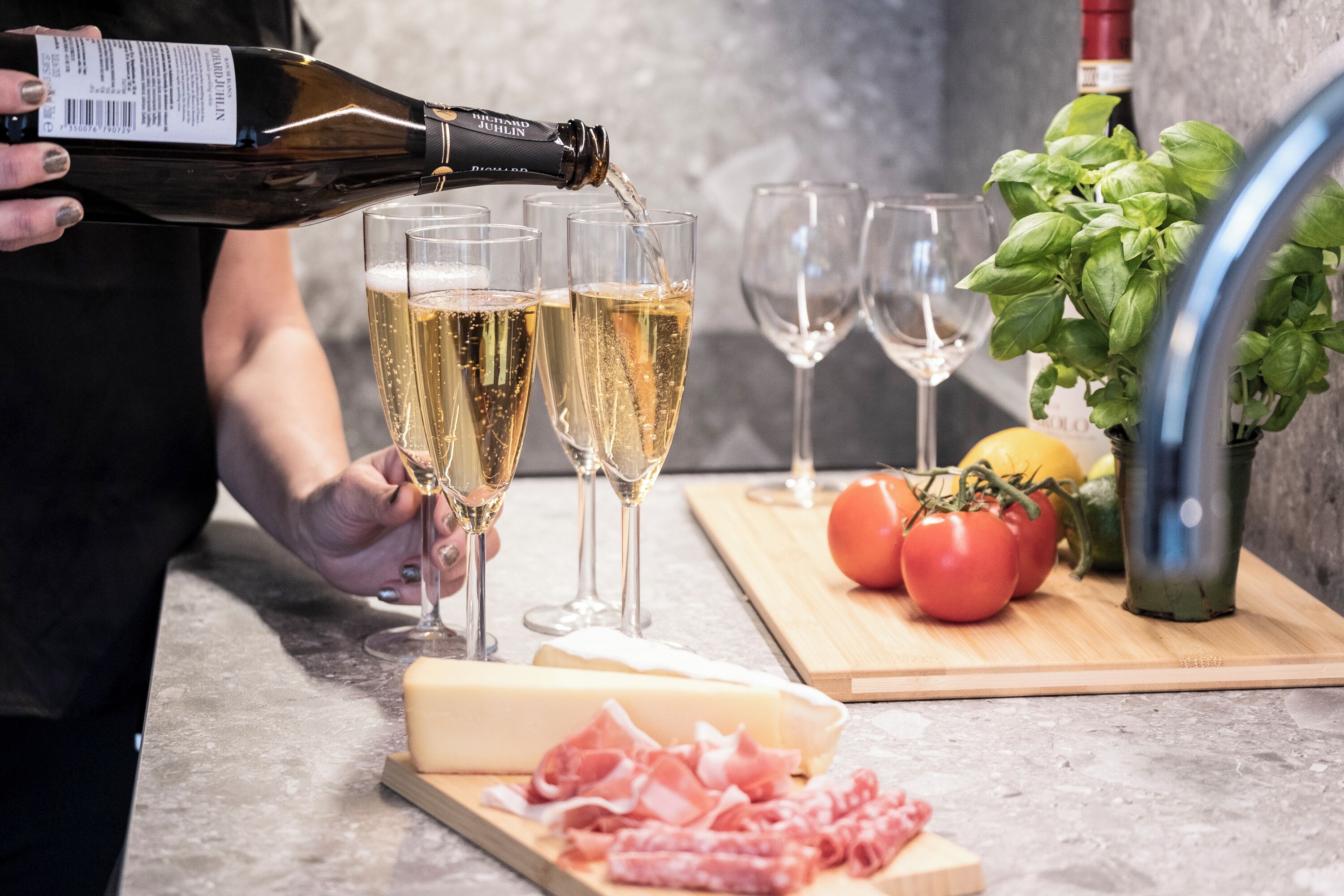 Eine Person gießt Champagner in Gläser, neben einem Käsebrett mit Tomaten und Basilikum auf einer Küchenarbeitsplatte.