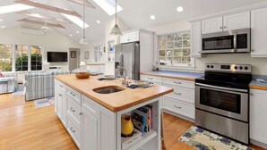 Eine moderne Küche mit weißen Schränken, einer Holz-Kochinsel mit Spüle, einem Edelstahl-Kühlschrank, einer Mikrowelle und einem Backofen. Fenster sorgen für natürliches Licht und bieten einen Blick ins Freie.