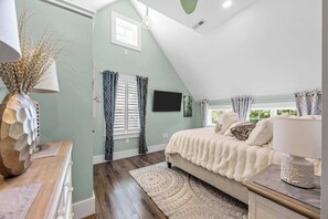Une chambre avec un plafond en pente, un lit recouvert d’une couverture blanche, un téléviseur fixé au mur et une fenêtre ornée de rideaux à motifs.