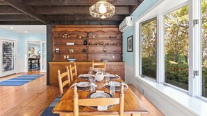 A dining area with a wooden table set for six, surrounded by chairs, with a wooden cabinet and shelves in the background. - Game Room, Walk to Two Landings! (Wellfleet)