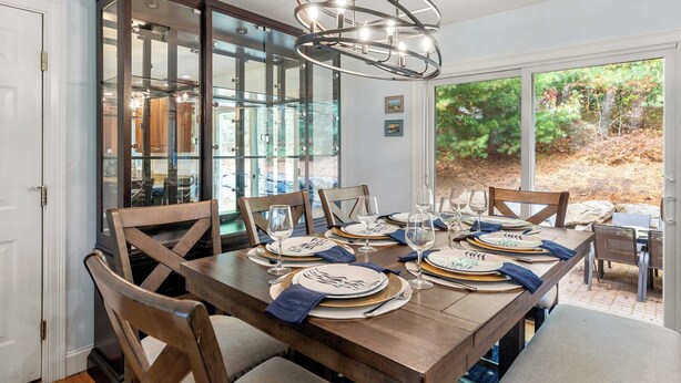Un comedor con una gran mesa de madera dispuesta para seis personas, rodeada de sillas de madera. Un candelabro moderno cuelga sobre la mesa, y la habitación tiene grandes puertas de vidrio que dan a un área exterior con vistas a árboles y un patio.