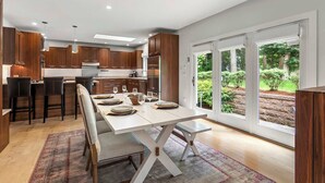 Een moderne keuken met houten kastjes, een eetgedeelte met een witte tafel en banken, en grote ramen met uitzicht op een tuin.