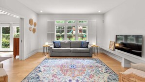 Een woonkamer met een grijze bank, een kleurrijk vloerkleed, een televisie en uitzicht op een tuin door grote ramen.