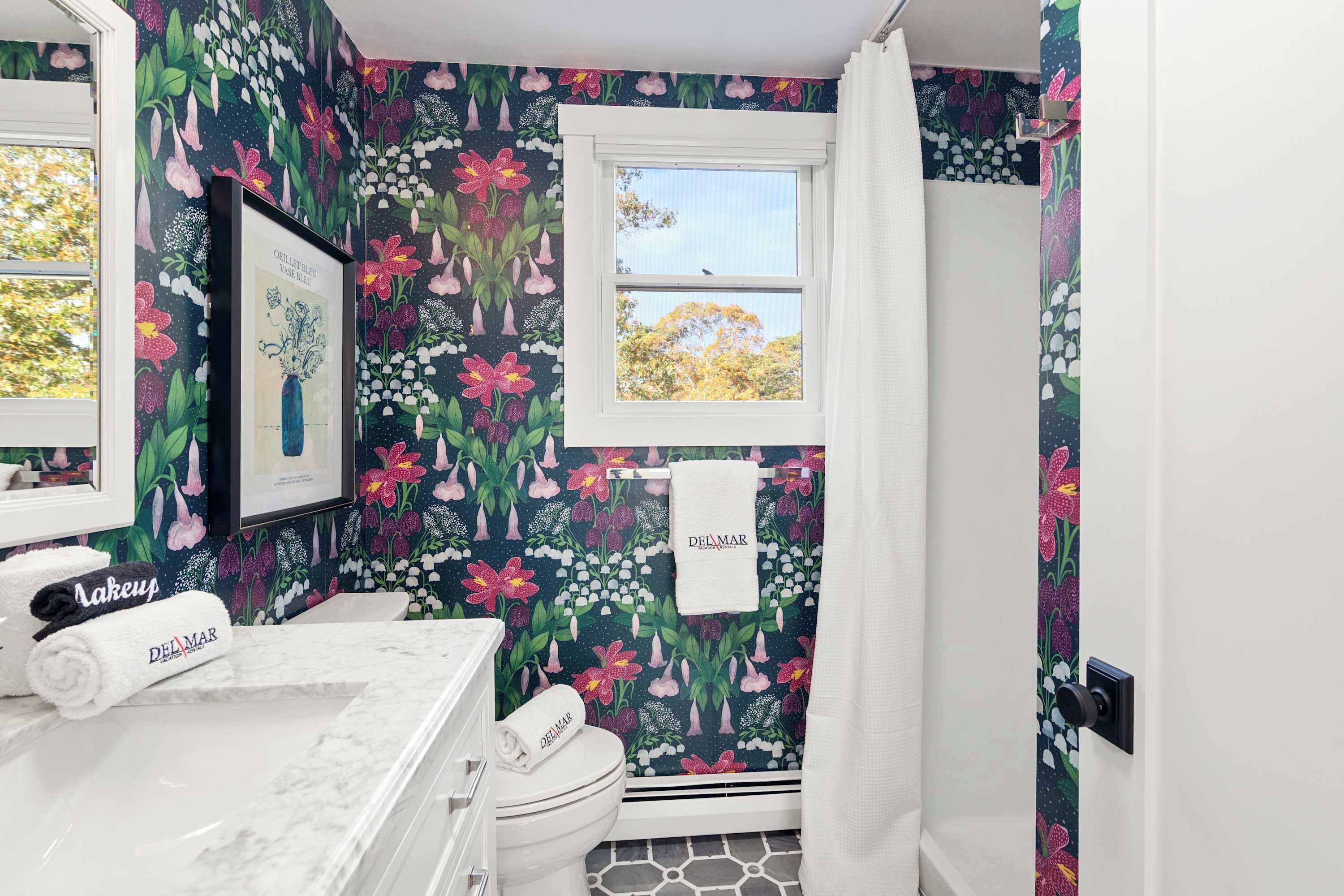 Un bany amb paper pintat floral, un vàter blanc, un lavabo amb taulell blanc i una finestra amb vista a arbres.