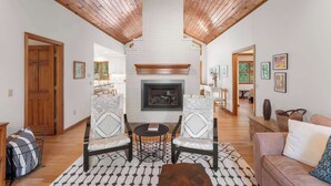 A spacious living room with a fireplace, wooden ceiling beams, and a view into the kitchen. - Game Room, Central AC, Short Drive to Beaches! (Eastham)