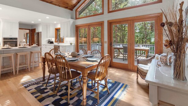 A spacious kitchen with white cabinets and a large island, a dining area with wooden chairs and a blue patterned rug, and large windows offering a view of trees. - Game Room, Central AC, Short Drive to Beaches! (Eastham)