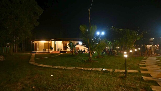 A modern building with a covered outdoor area, surrounded by a well-maintained lawn and palm trees. - JUNGLE FARMS BY MAARK INN (Kanakapura)