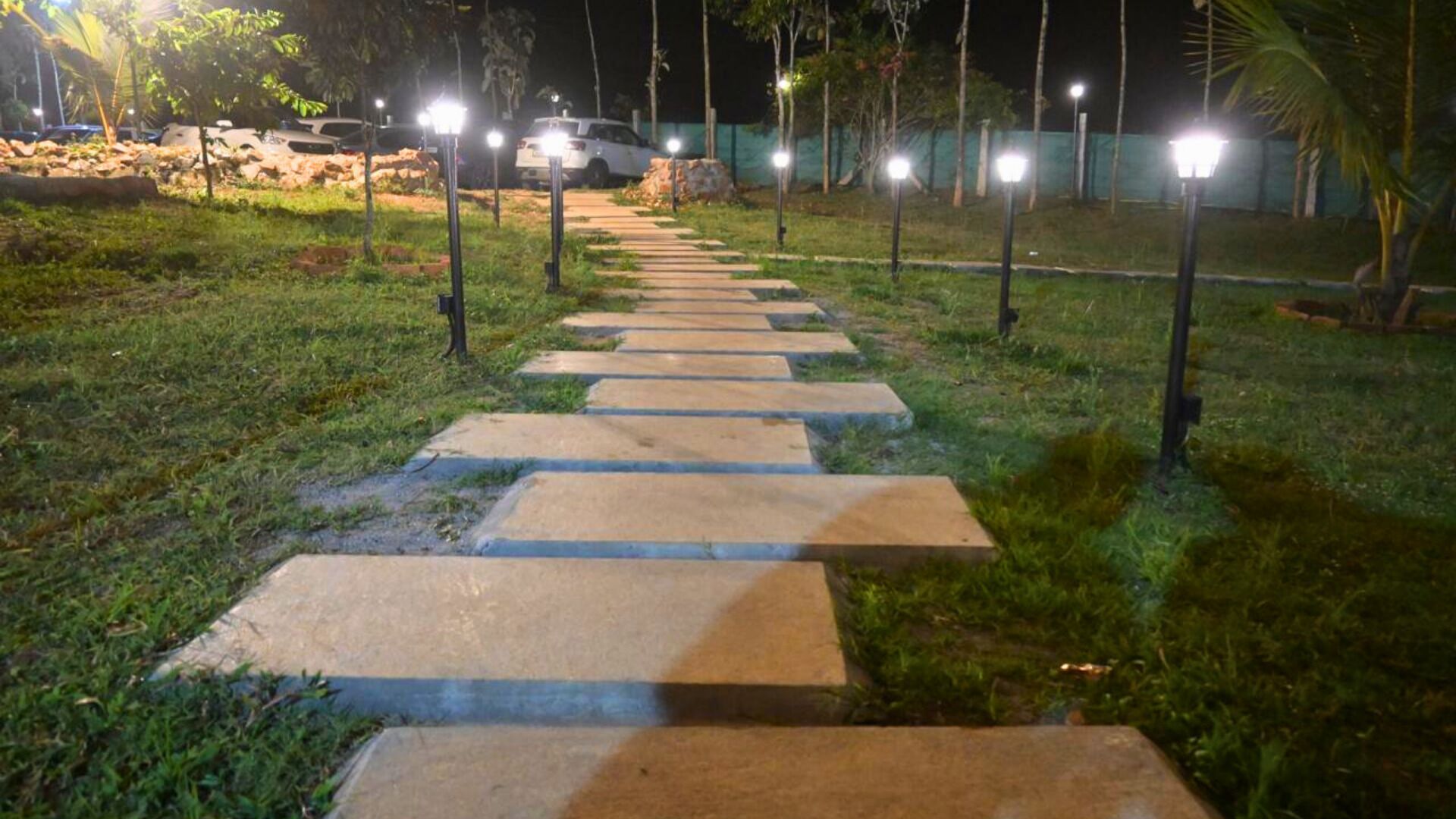 Un sendero nocturno con losas de piedra y farolas, rodeado de césped y árboles.