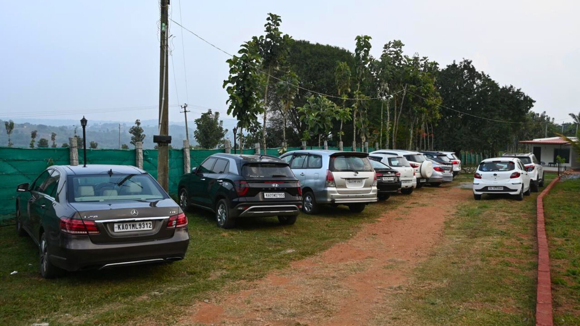 A parking area with multiple cars parked along a dirt path, surrounded by greenery and a fence.