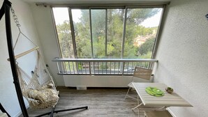 A room with a hammock, a table with a green mat, and a view of trees and buildings through large windows. - Bright studio a few steps from the beach (Saint-Mandrier-sur-Mer)