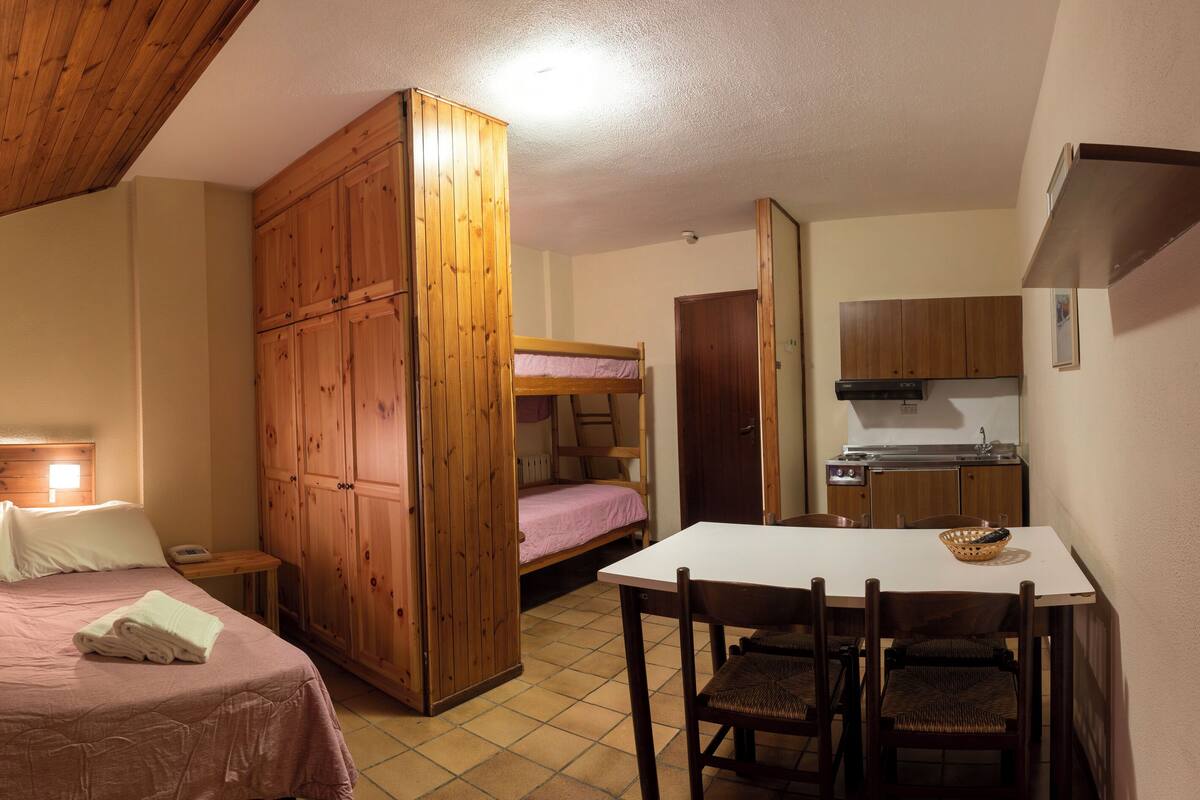 A bedroom with a bunk bed, a wooden wardrobe, a bedside table with a lamp, and a small kitchen area with a sink and cabinets.