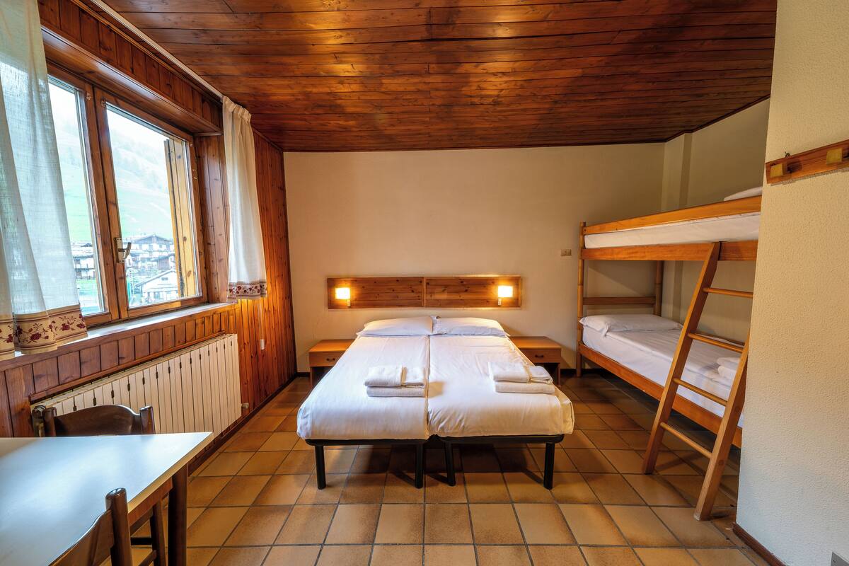 A room with a wooden ceiling, a large bed with two pillows, a bunk bed, a table with chairs, and a window with curtains.