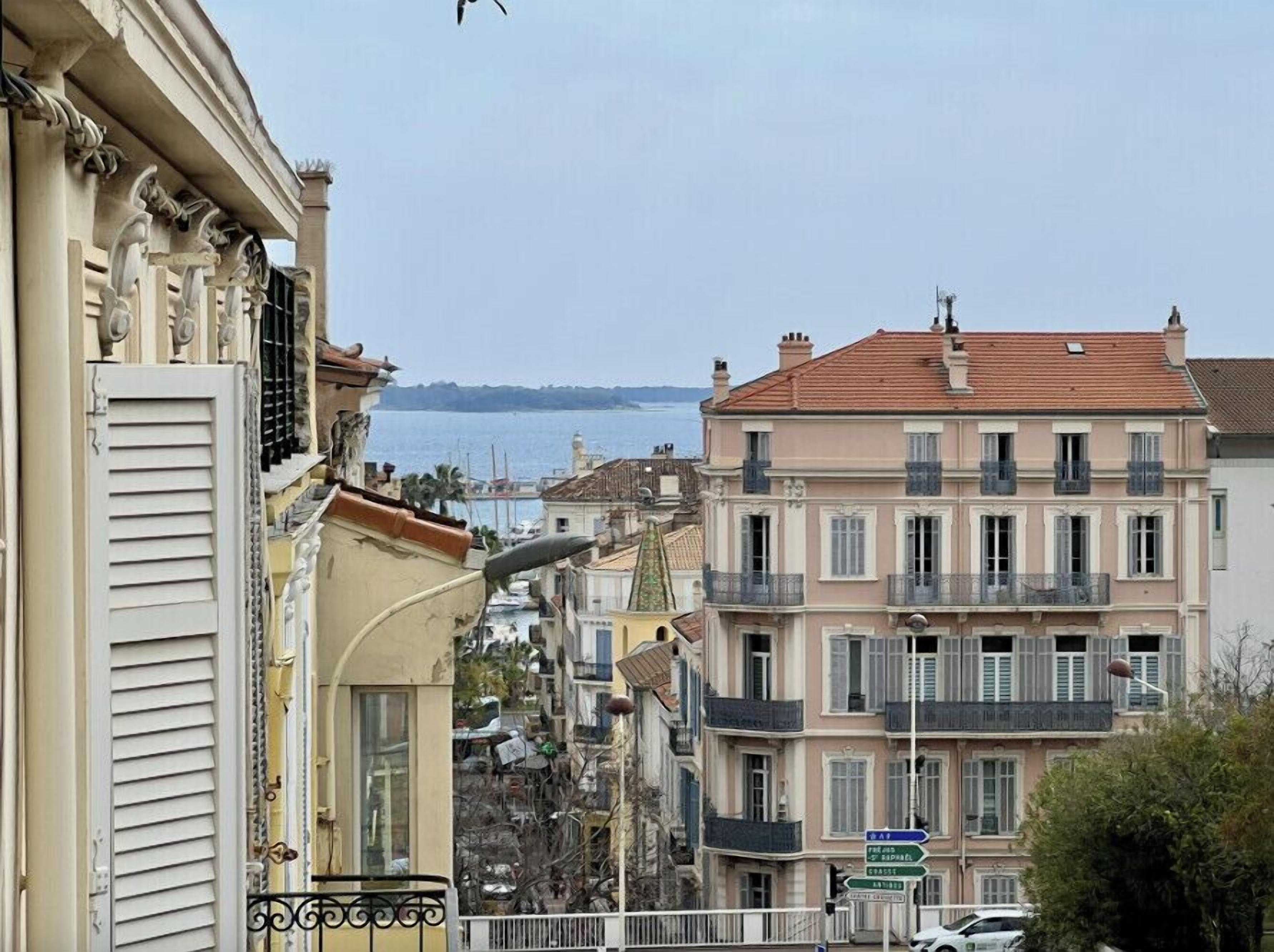 A coastal cityscape with multi-story buildings, a clear view of the sea, and a street with a directional sign.