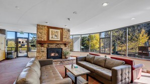A spacious lounge area with leather sofas, a stone fireplace, and large windows offering a view of trees and a building. - Spacious ski-in/ski-out Studio, 1-Bath Prime Location to Ski Slopes! (Breckenridge)