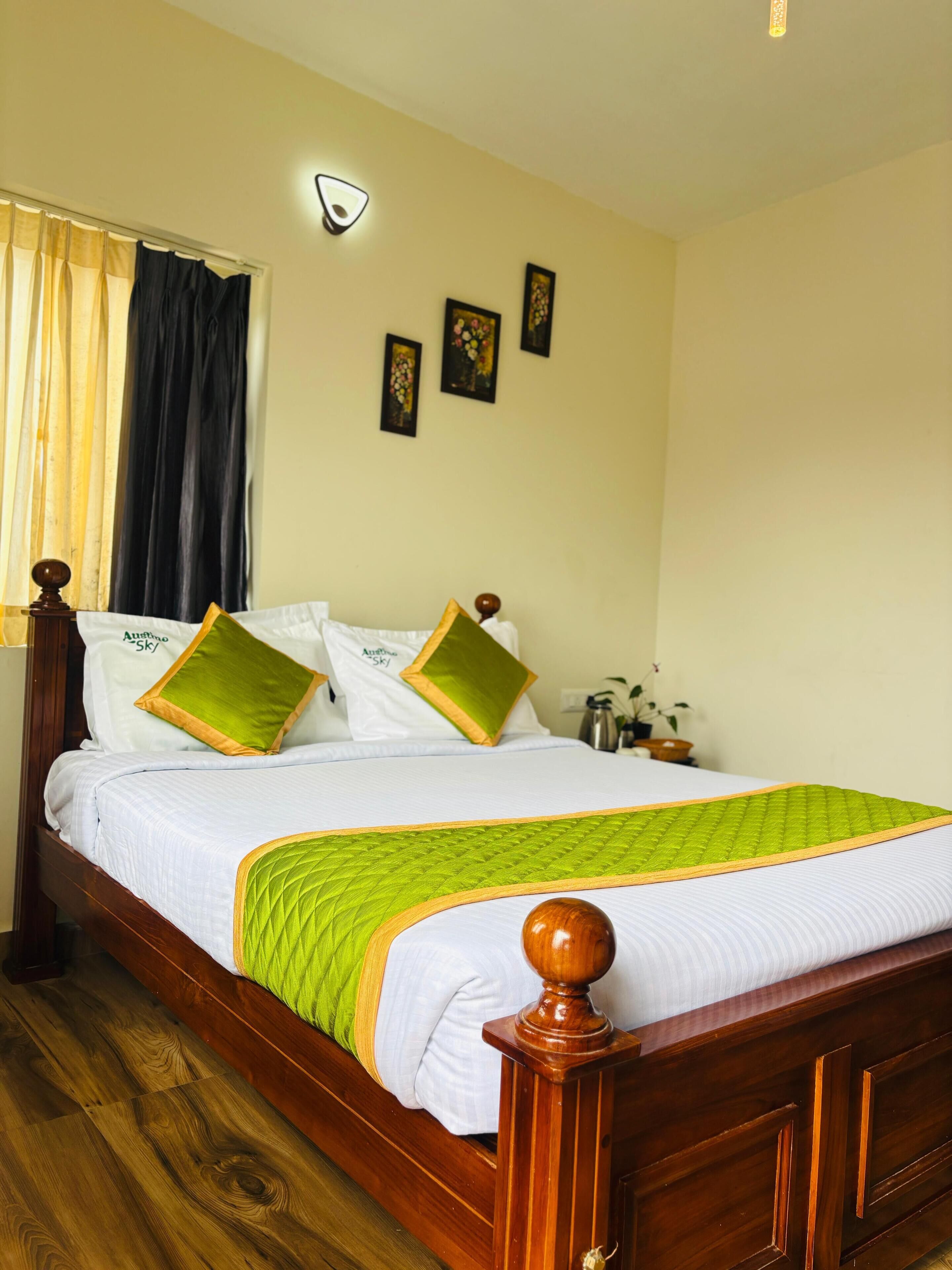 A neatly made bed with a wooden headboard, white bedding, and green and gold pillows. There are three framed pictures on the wall, a bedside table with a lamp, and a window with curtains.