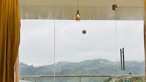Une chambre d’hôtel dotée d’une grande fenêtre, de rideaux, d’un luminaire suspendu et offrant une vue sur un paysage vallonné.