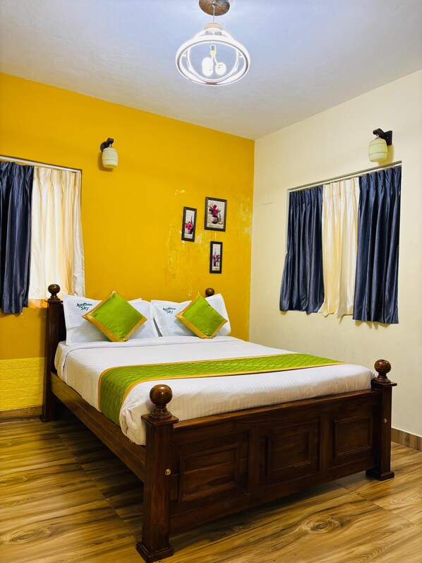 Een net opgemaakt bed met een houten frame, wit beddengoed en groene kussens. Er hangen drie ingelijste foto's aan de muur, een plafondlamp en een raam met blauwe gordijnen.