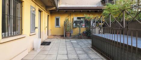 Un patio estrecho y pavimentado con un edificio amarillo, puertas metálicas y plantas en macetas.