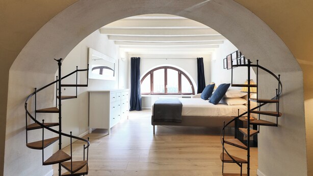 A bedroom with a curved staircase, a bed with blue pillows, and a large arched window. - Brera Apartments in Gae Aulenti (Milan)