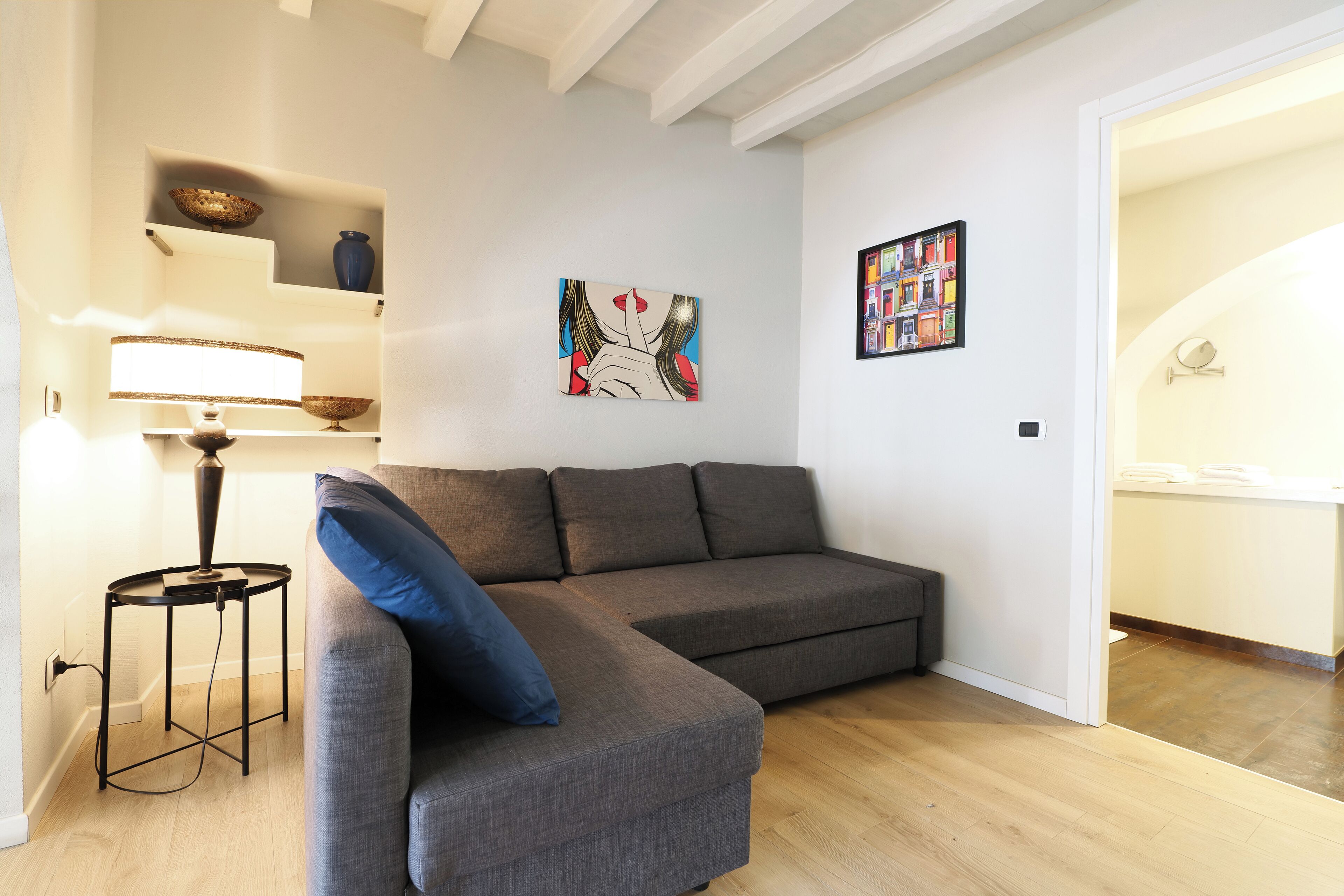 A modern living room with a grey sectional sofa, a small black side table, and a floor lamp. There are two wall-mounted shelves with decorative items and two framed artworks.