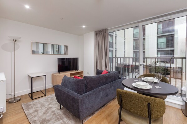 A modern living room with a grey sofa, a flat-screen TV, a dining table with chairs, and a large window with a view of buildings. - Olive & Stone - Studio Apartment, Sleeps 2 (Bristol)