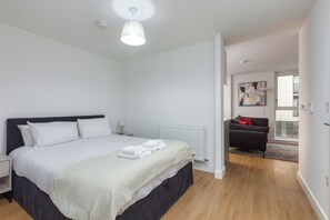 A modern bedroom with a large bed, bedside tables, a radiator, and a view to a balcony with a sofa. - Olive & Stone - Studio Apartment, Sleeps 2 (Bristol)