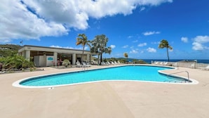 A pool area with a clear blue sky, palm trees, and a building with a covered seating area. - Generator plus Beach, Pool, Golf, Tennis, Pickleball @ The Reef East End STX (Christiansted)