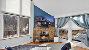 A cozy living room with a fireplace, large windows, and a view of a house. - Dog-Friendly Chalet in The Woods Resort w/ Fire Pit (Hedgesville)