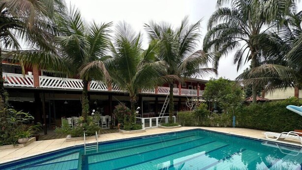 Piscina com água azul cristalina, rodeada por palmeiras e um edifício com telhado vermelho e grades brancas.