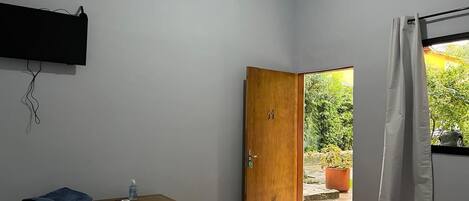 A bedroom with a bed, a wooden desk, a wall-mounted air conditioner, and a door leading to a balcony with potted plants.