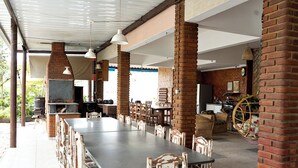 Uma área de refeições ao ar livre coberta, com pilares de tijolo, uma mesa longa e cadeiras rústicas.