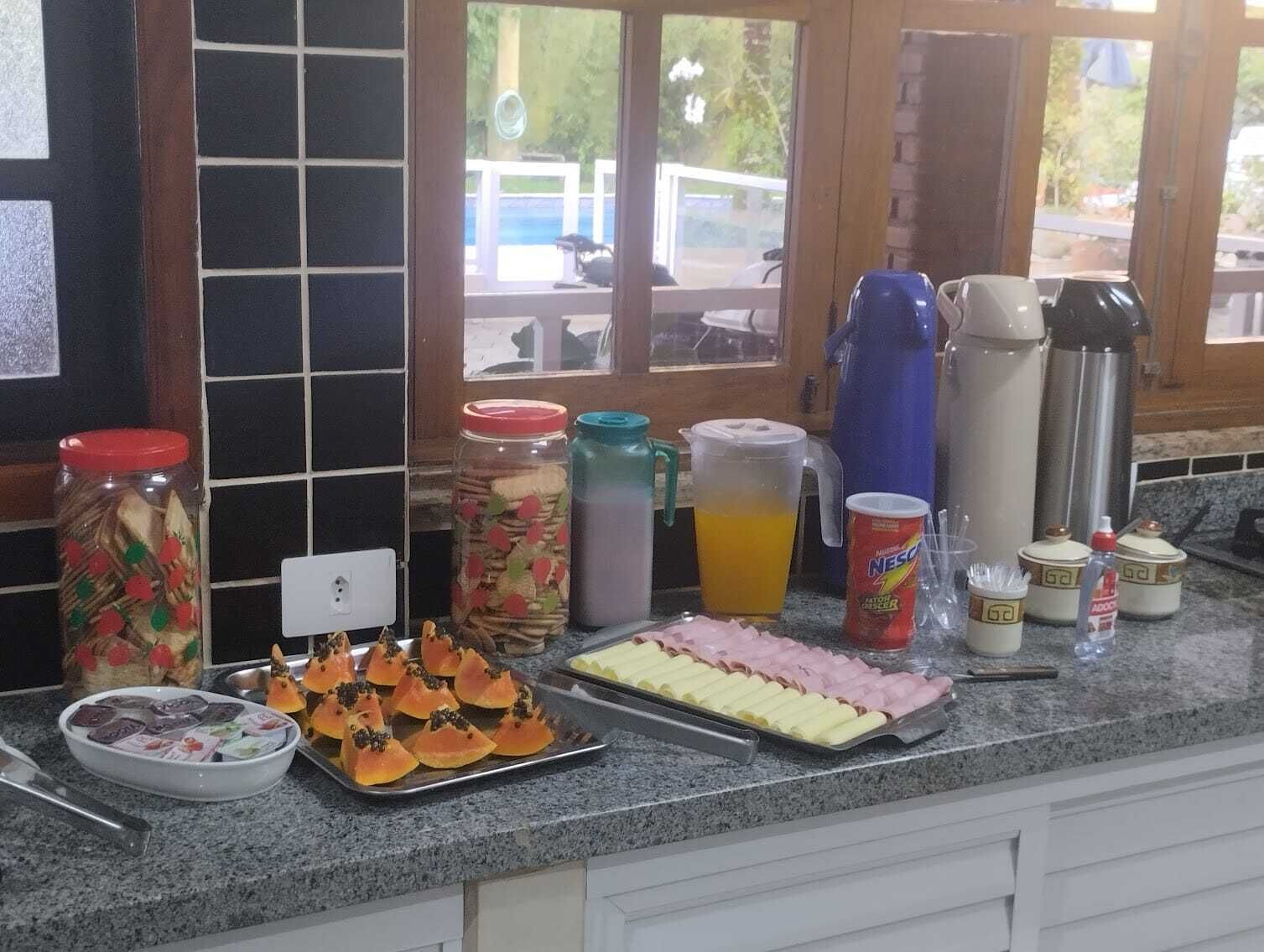 A kitchen counter with various food items and condiments, including jars of pickled vegetables, a tray of sliced fruits, a pitcher of orange juice, and a can of Nescafé instant coffee.