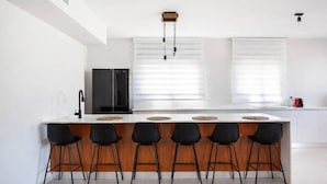 A modern kitchen with a central island, black bar stools, a black refrigerator, and a window with blinds. - Beachside 3BR | Huge Patio| Mamad (Bat Yam)