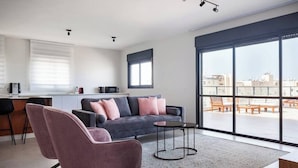 A modern living room with a grey sofa, pink chairs, a coffee table, and large windows offering a city view. - Beachside 3BR | Huge Patio| Mamad (Bat Yam)
