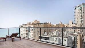 A balcony with a wooden floor, a lounge chair, and a view of a cityscape with buildings and a body of water. - Beachside 3BR | Huge Patio| Mamad (Bat Yam)