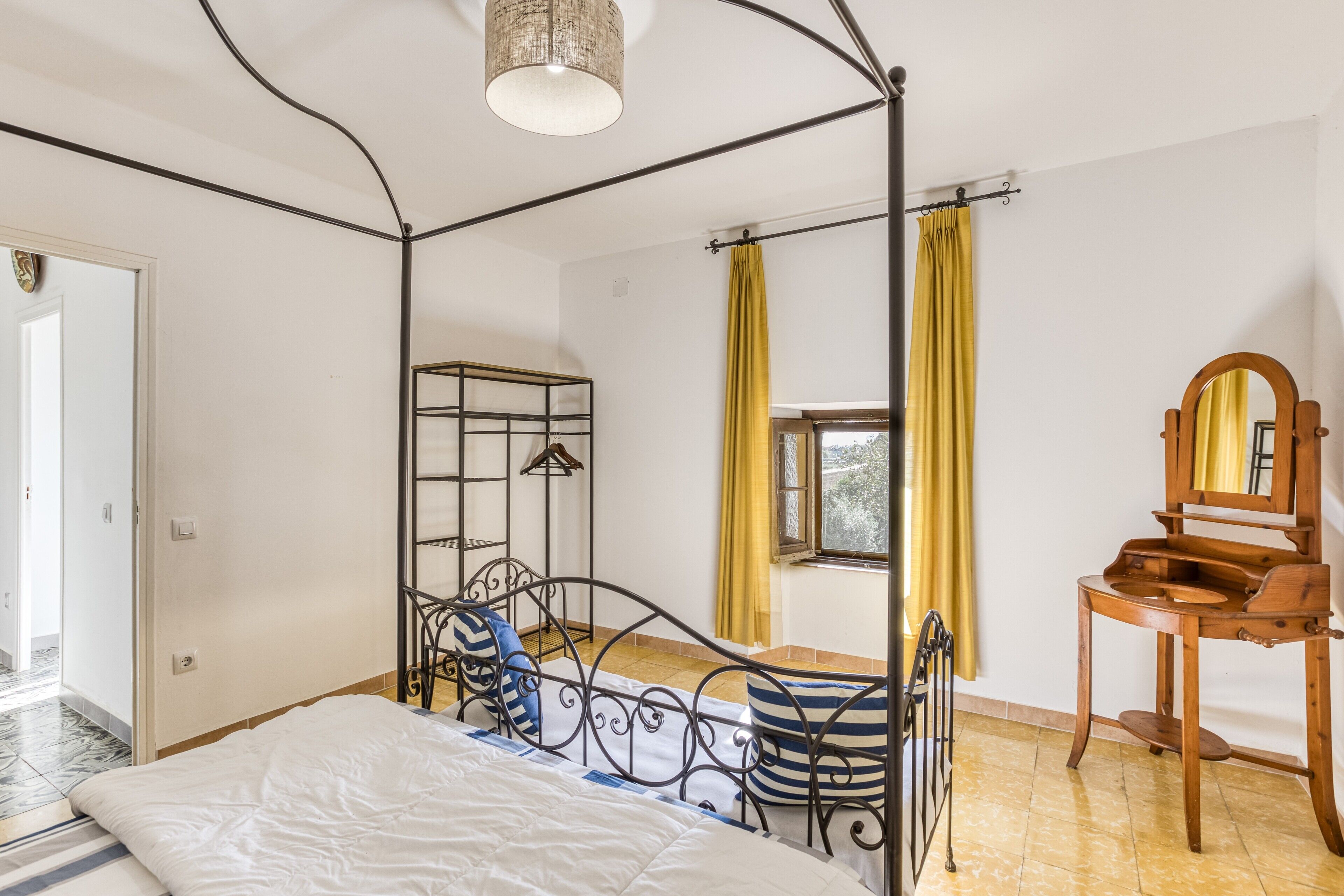 Une pièce avec un lit en métal, une chaise en bois, un miroir et une fenêtre ornée de rideaux jaunes.