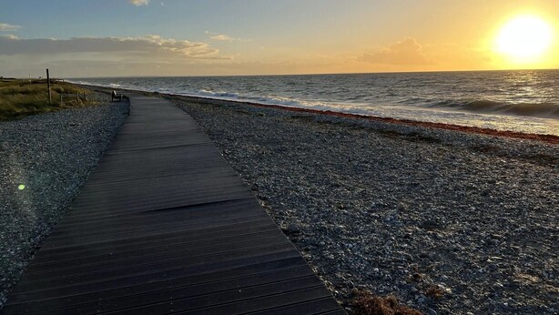 Um caminho à beira-mar que leva ao oceano, com um pôr do sol ao fundo.