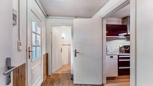 Um corredor com piso de madeira, porta branca e área de cozinha com armários e eletrodomésticos.