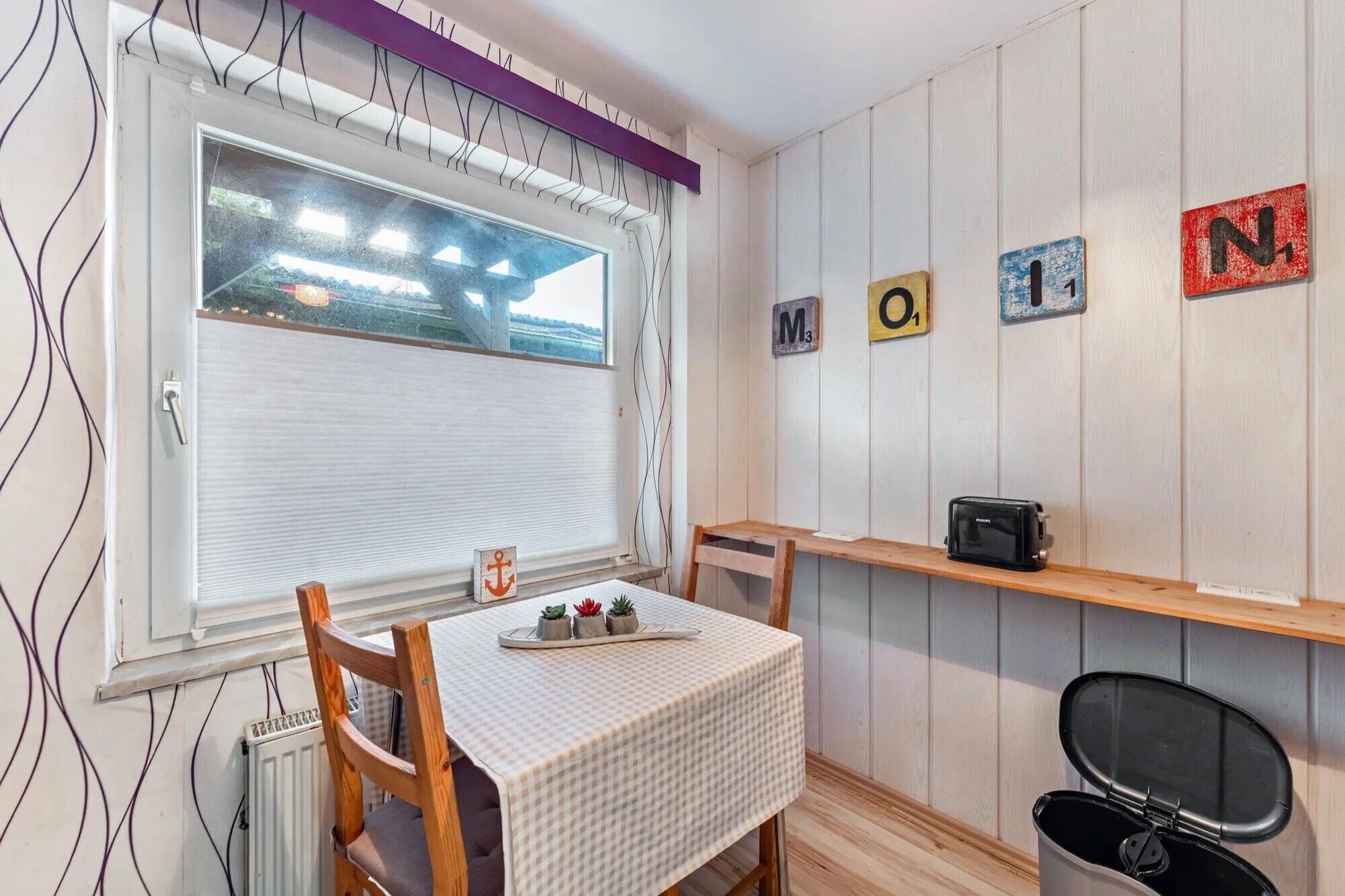 Una pequeña cocina con una ventana, una mesa con mantel a cuadros, una silla, un estante con un tostador y un cubo de basura.