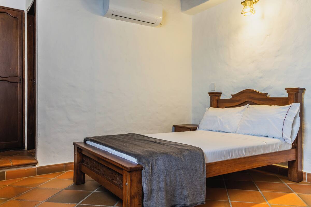 A bedroom with a wooden bed, a nightstand, a bench, a wall-mounted air conditioner, and a light fixture.