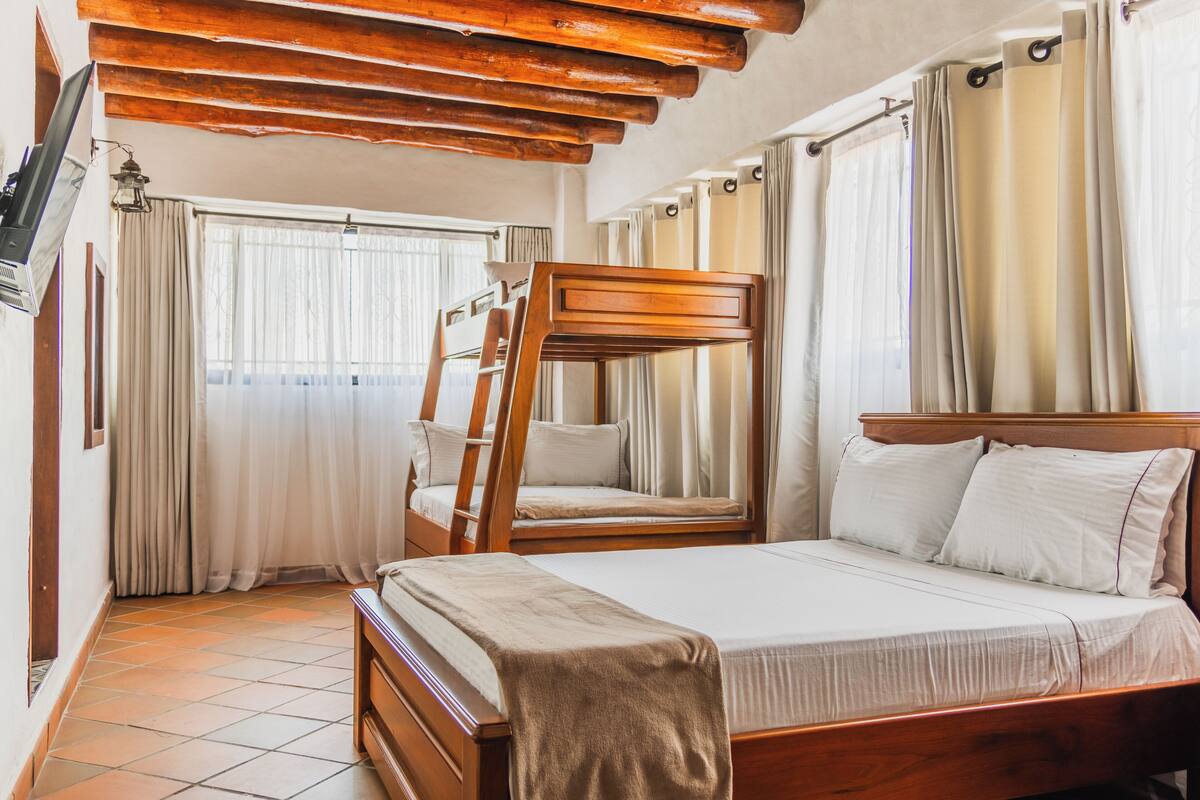 A bedroom with a bunk bed, a wooden headboard, a large bed with white linens, and a window with curtains.