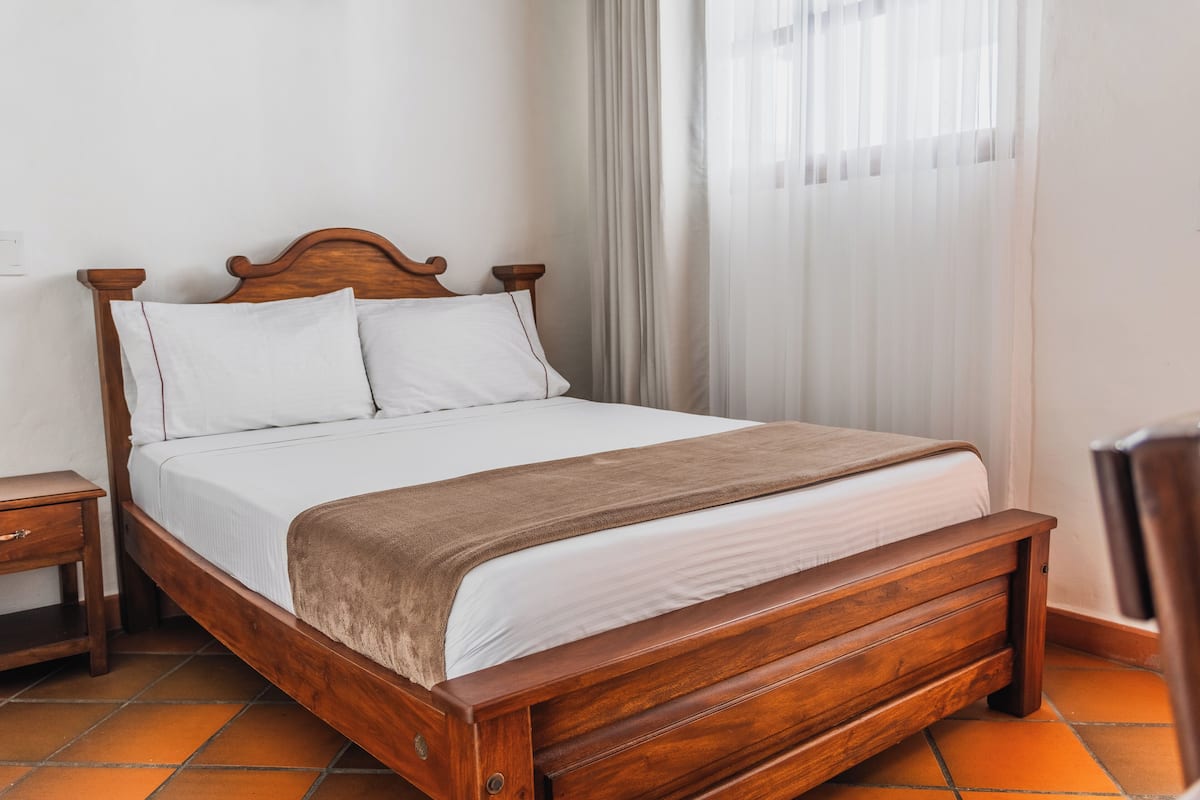 A wooden bed with white linens and a brown throw blanket in a room with a tiled floor and a window with sheer curtains.