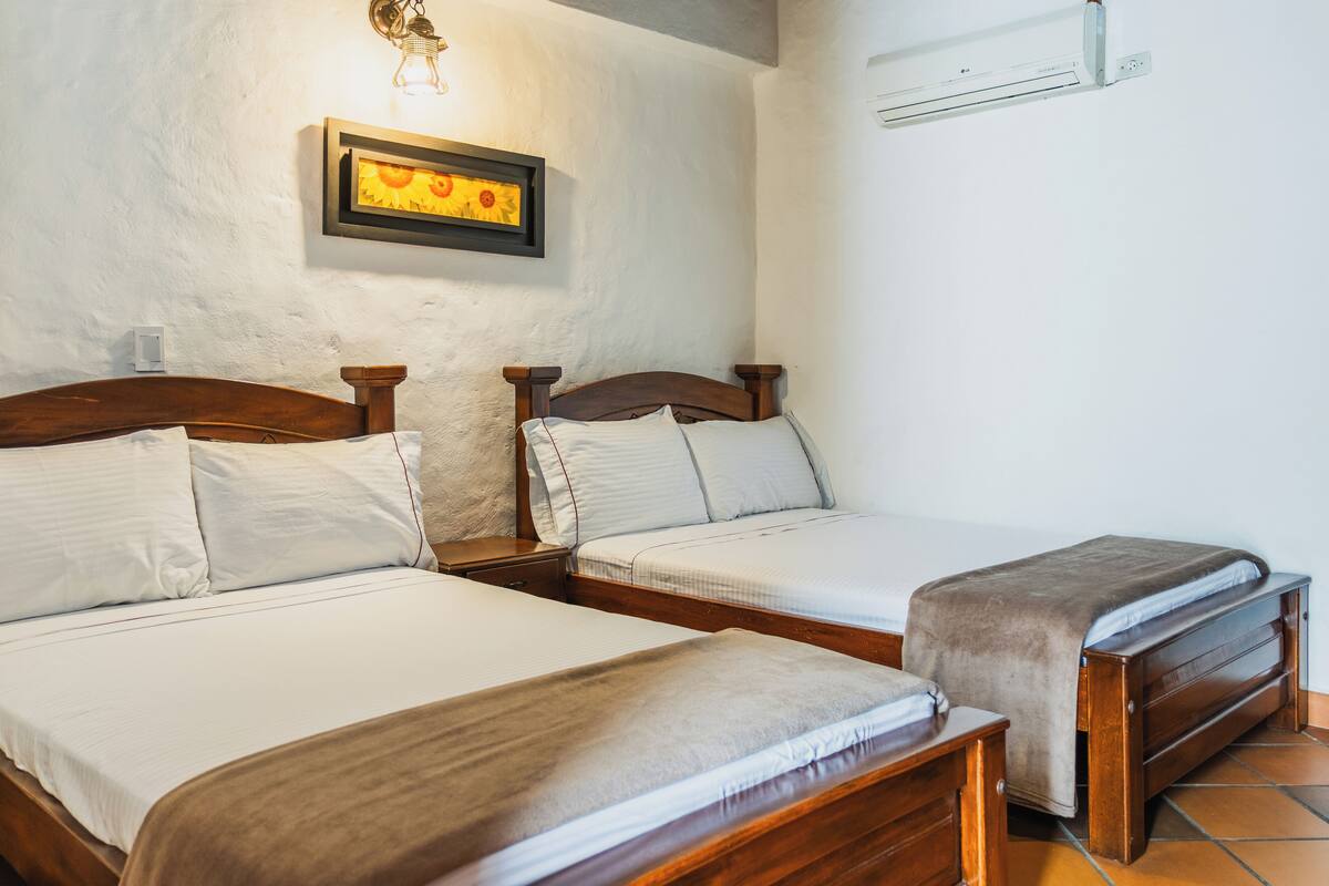 A bedroom with two wooden beds, white bedding, a wall-mounted light fixture, and an air conditioning unit.