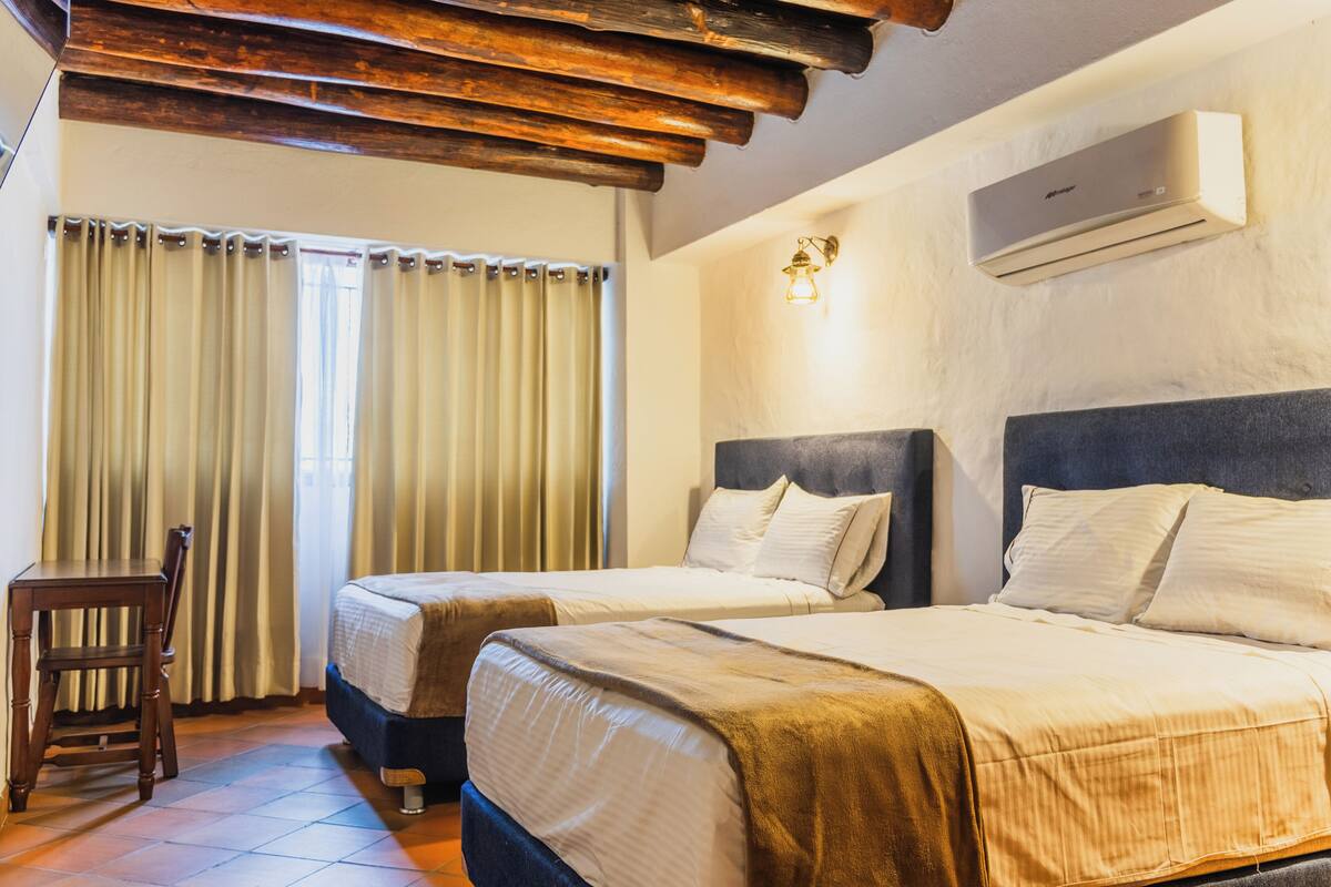 A hotel room with two beds, a wooden ceiling, an air conditioner, and a desk with a chair.