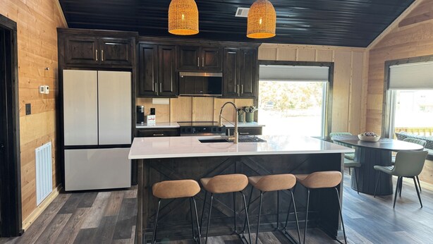 Een moderne keuken met donkere houten kastjes, een roestvrijstalen koelkast, een groot eiland met barstoelen, hanglampen en een uitzicht door een groot raam.