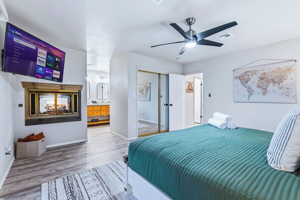 A modern bedroom with a bed, a ceiling fan, a mounted TV displaying streaming services, a world map on the wall, and a view into the bathroom.