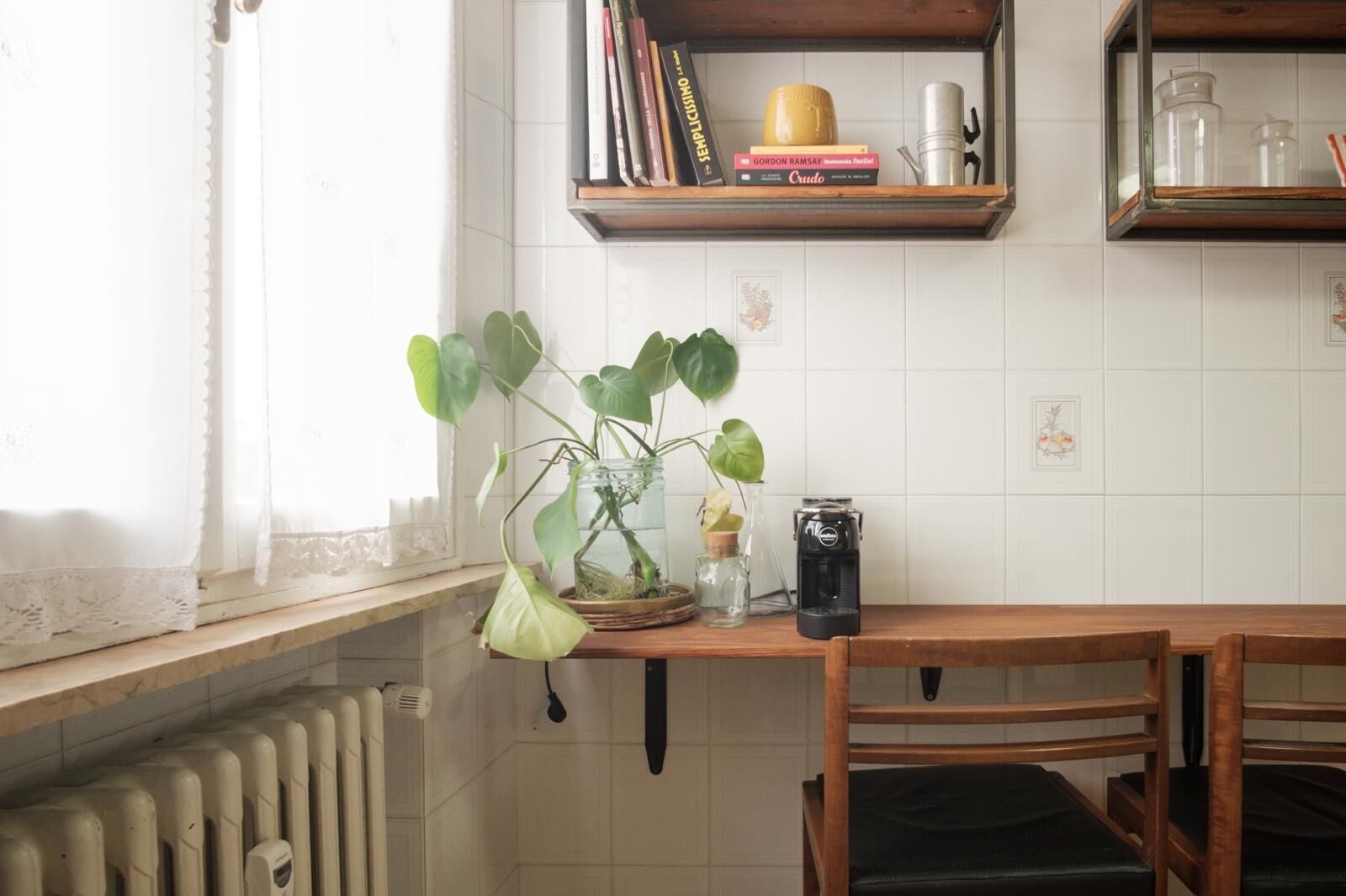 A kitchen with a wooden table, chairs, a potted plant, a coffee maker, and a shelf with books and kitchenware.