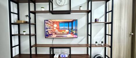 A living room with a TV mounted on a black-framed wall, surrounded by wooden shelves, a clock, and a fan.