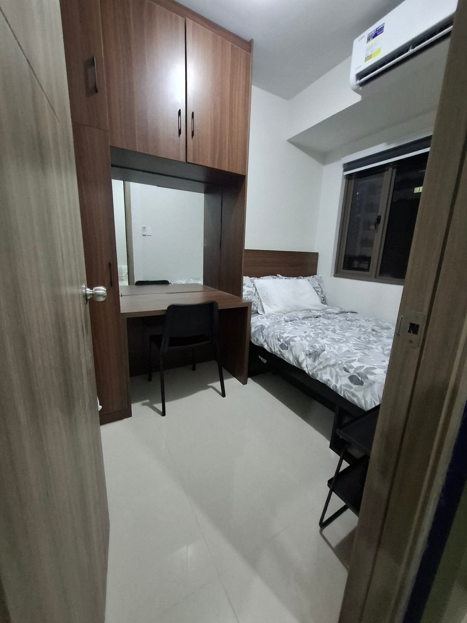 A compact bedroom with a wooden wardrobe, a desk, a bed with a patterned bedspread, and an air conditioning unit.